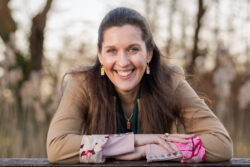 A outdoor business headshot taken of a woman leaning forward and smiling directly at the camera.