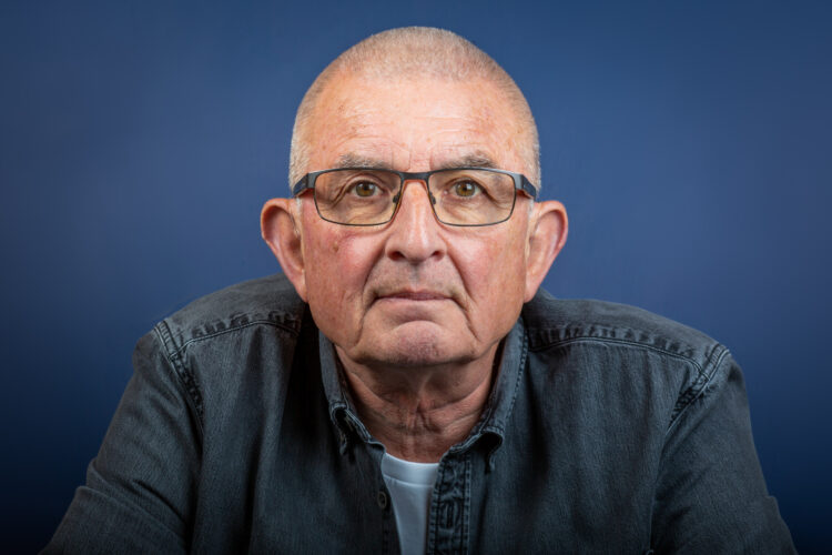 A business headshot of a man sat square on to the camera, gazing intently at the viewer. Blue background.
