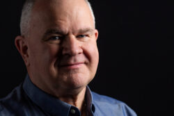 A dramatic close-up business headshot of a man standing against a black background and with strong shadows
