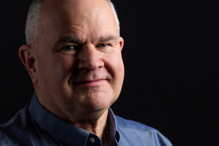 A dramatic close-up business headshot of a man standing against a black background and with strong shadows
