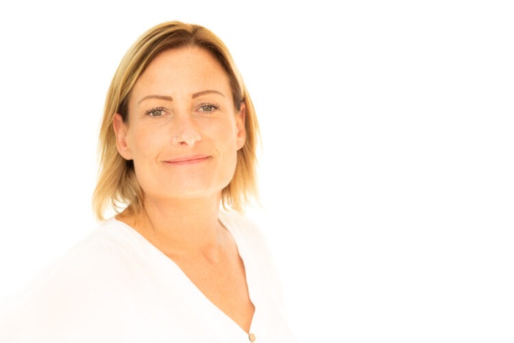 A business headshot of a woman wearing a white top standing in front of a bright white background