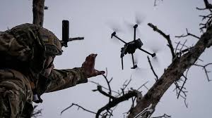 A soldier in uniform, wearing a helmet and tactical gear, kneels down to retrieve a quadcopter drone from the sky after a successful mission.