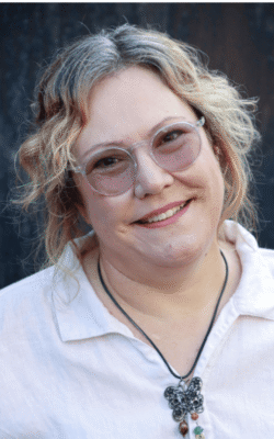 Professional headshot of Melanie Carr, founder of Diverse Perspectives Ltd. Melanie is a white woman with curly blonde and grey hair, wearing round clear-framed glasses, a white linen shirt, and a black cord necklace with a decorative butterfly pendant. She is smiling warmly at the camera against a dark blurred background.