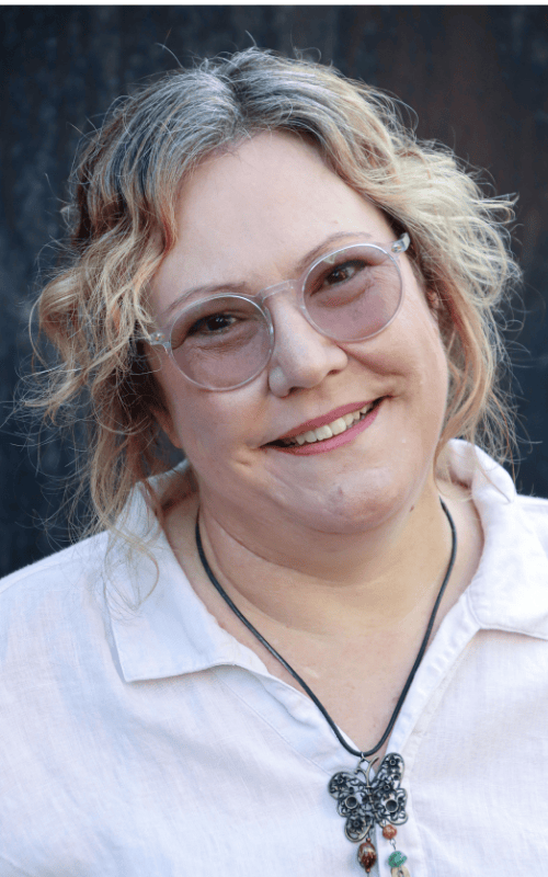 Professional headshot of Melanie Carr, founder of Diverse Perspectives Ltd. Melanie is a white woman with curly blonde and grey hair, wearing round clear-framed glasses, a white linen shirt, and a black cord necklace with a decorative butterfly pendant. She is smiling warmly at the camera against a dark blurred background.