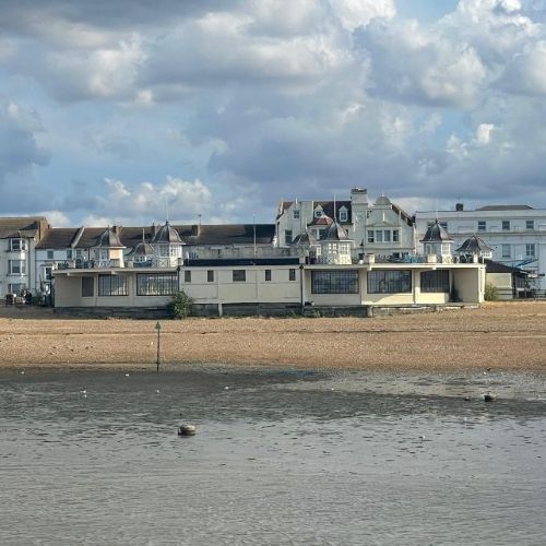 A seaside building with large windows sits on a sandy beach at low tide, with older waterfront houses and a cloudy sky in the background.