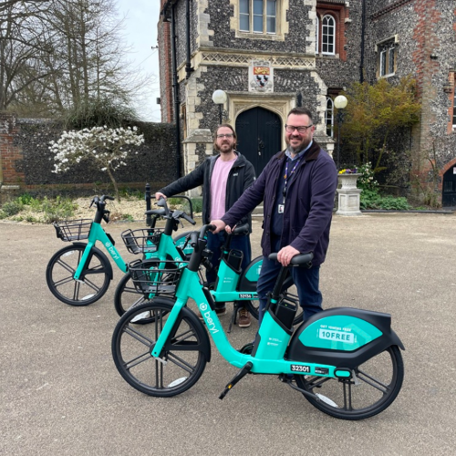 Two people standing outdoors with turquoise electric bikes in front of a historic brick and flint building. Each person is holding a bike upright, with two additional bikes beside them. A small blossoming tree, other plants and an historic building are visible in the background