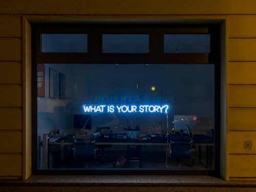 A darkened office window, showing a blue neon sign with the text, What's your story?
