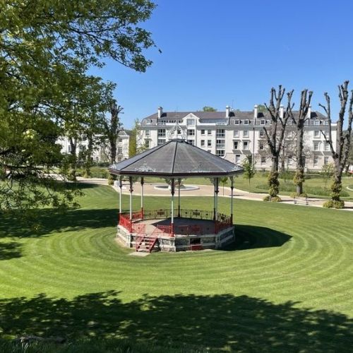 Bandstand with a grey roof and red railings set on a circular, closely mown lawn, with a large white multi‑storey building in the background and trees framing the scene under a clear blue sky.