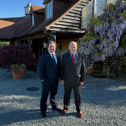 Two people wearing dark suits and ties stand side by side on a gravel driveway in front of a building with wooden cladding and a sloped roof. A clock tower rises from the roof on the left. A large flowering vine with purple blossoms covers part of the wall on the right, and potted plants and shrubs line the building. Bright sunlight casts clear shadows across the scene.