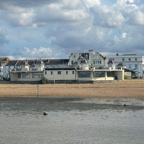 Seafront bandstand building viewed from across a pebble beach and tidal shoreline, with residential buildings and cloudy skies in the background.