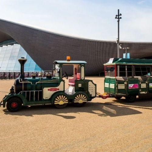 Small green road train with a passenger carriage parked outside a modern building with a curved roof.