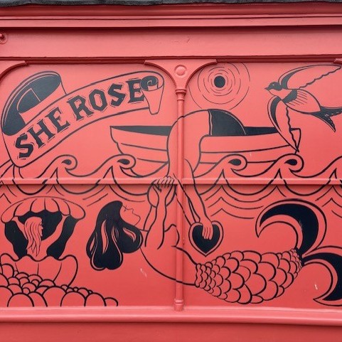 Coral-pink mural featuring illustrated sea-themed artwork, including a banner reading 'SHE ROSE', ocean waves, a bird in flight, shells, and a mermaid tail