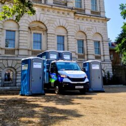 A branded euroloo service truck parked outside a grand historic building, flanked by portable toilets on either side with additional units loaded on the truck bed.