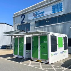 Two white and green euroloo ecowelfare mobile units with extended solar panel awnings parked outside a commercial warehouse building.