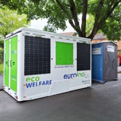 A white and green euroloo ecowelfare mobile unit with side solar panels parked on a paved road next to a single portable toilet under trees.