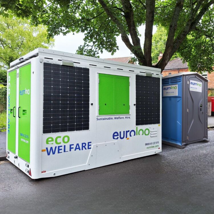 A white and green euroloo ecowelfare mobile unit with side solar panels parked on a paved road next to a single portable toilet under trees.