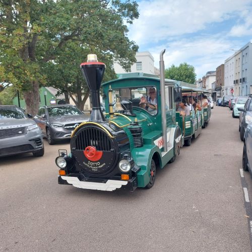 Green land train carrying passengers drives along a town street lined with parked cars, trees and buildings
